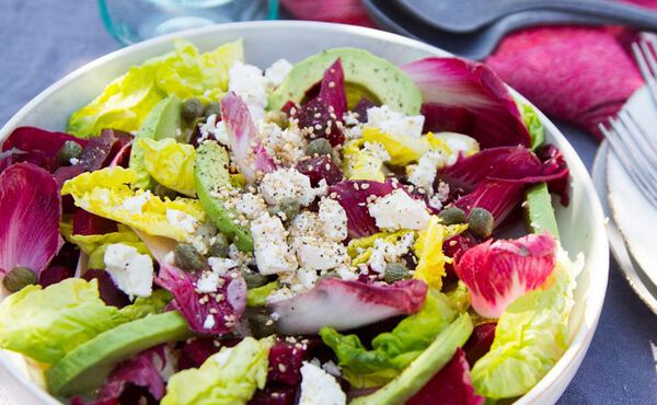 Bowl of healthy dinner salad with avocado, feta, capers and fresh greens on a table.