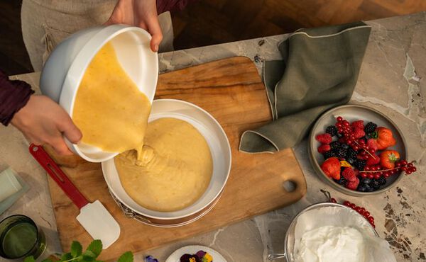 A person pouring yellow cake batter from a mixing bowl into a round baking tin on a wooden board, surrounded by fresh berries, whipped cream, a spatula, herbs, and edible flowers on a marble countertop.