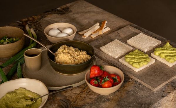 Ingredientes para preparar sandos japoneses, un tipo de sándwich, están dispuestos sobre una encimera de mármol: rebanadas de pan blanco untadas con crema de matcha, un bol de ensalada de huevo, huevos enteros, cebolletas, un bol de fresas frescas y los bordes del pan recortados.