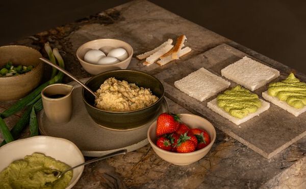 Les ingrédients des sandos japonais, un type de sandwich, sont disposés sur un plan de travail en marbre, avec des tranches de pain tartinées de crème de matcha vert, un bol de salade d’œufs, des œufs entiers, des oignons verts, un bol de fraises fraîches et les croûtes coupées du pain.