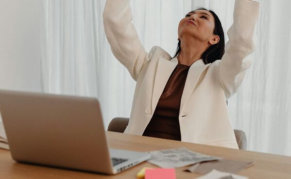 Woman stretching at her desk with eyes closed and arms raised, taking a mindful pause during the workday.