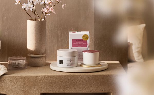 The Ritual of Sakura refillable body cream and scented candle refill shown on a bedside table next to a cherry blossom plant.