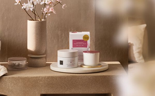 The Ritual of Sakura Refill Body Cream og Scrented Candle Refill vist på et natbord ved siden af en kirsebærblomst.