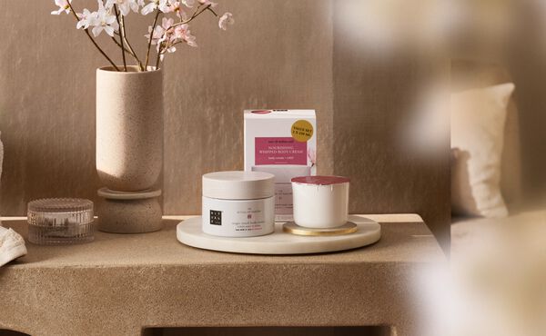 Crème pour le corps rechargeable et recharge pour bougie parfumée The Ritual of Sakura, disposées sur une table de nuit à côté d’une branche de fleurs de cerisier.