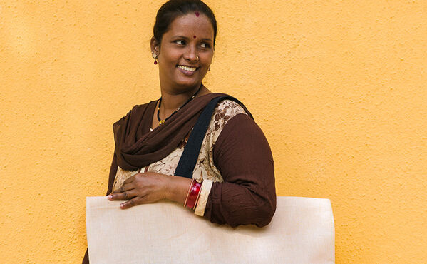 Una mujer de la comunidad Tiny Miracles sonríe mientras lleva una bolsa hecha a mano y posa delante de un muro de color amarillo.