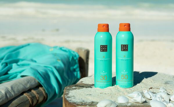 To flasker af The Ritual of Karma SPF 30 og SPF 50 Sunscreen Spray på stranden ved siden af muslingeskaller og en turkis klud.