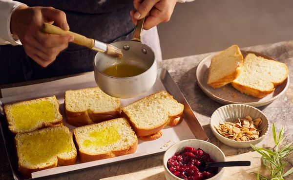 A person brushing slices of brioche with melted butter for a festive brunch, with cranberry sauce and flaked almonds nearby.