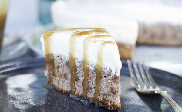 En bit banankaka med cheesecakeöverdrag och kolasås på en mörkblå tallrik med en gaffel.