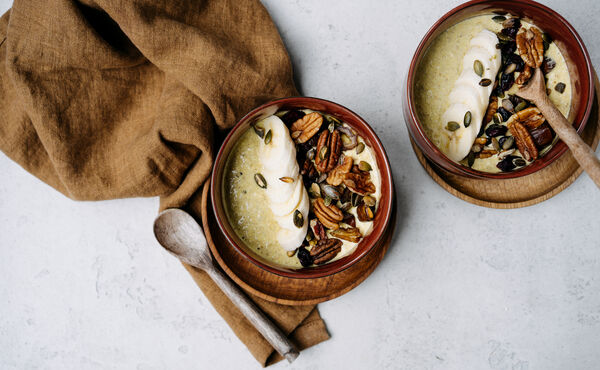 Two smoothie bowls topped with banana, pecans and seeds, served with wooden spoons on a light table.