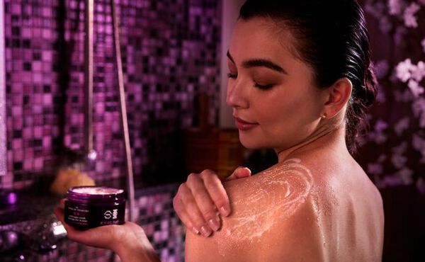 Woman applying The Ritual of Yozakura body scrub in a purple-lit bathroom, enjoying a relaxing skincare moment.