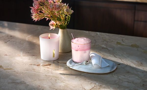 The Ritual of Sakura inspired mocktail with pink foam, next to a matching scented candle and vase of flowers.