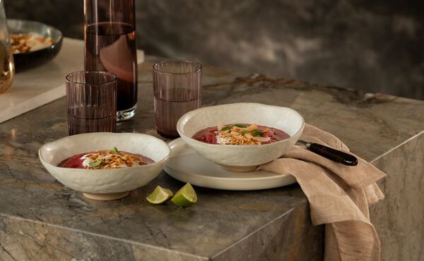 Miso-Brühe mit Pilzen: Schale mit Pilz-Miso-Suppe mit Nudeln, garniert mit Erbsen und Pilzen, serviert auf einem Holztablett mit Stäbchen und einem schwarzen Löffel, daneben ein Glas Wasser und eine braune Vase mit grünen Blumen auf einer Arbeitsplatte aus Marmor.