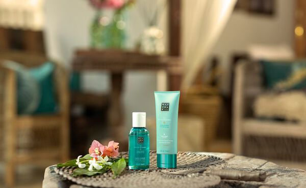 Zwei The Ritual of Karma Travel-Size Mini-Produkte in einer entspannten Sommerurlaubsatmosphäre auf einem Tisch mit Blumen.