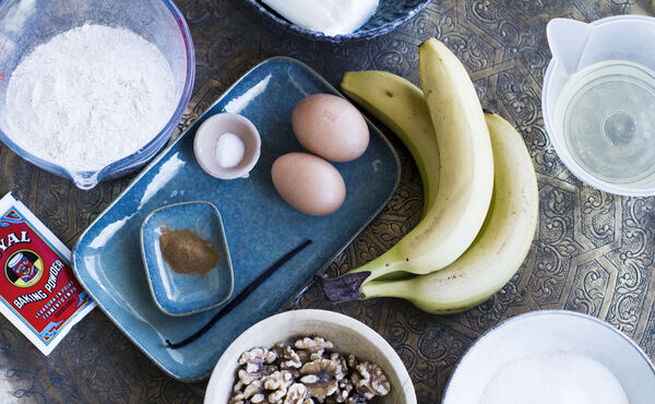 Ingredienser till en banankaka – ägg, banan, mjöl, valnötter och kryddor – står på ett bord, redo att användas.