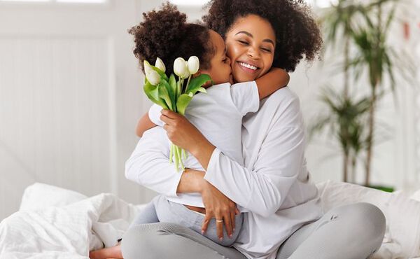 Uśmiechnięta kobieta siedzi na łóżku i przytula dziecko, które trzyma mały bukiet białych tulipanów, w jasnym, przytulnym pomieszczeniu oświetlonym światłem dziennym.