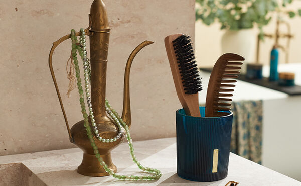 Photophore bleu avec des reliefs décoratifs utilisé pour ranger un peigne en bois et une brosse, disposé à côté d’une carafe en laiton avec des perles.