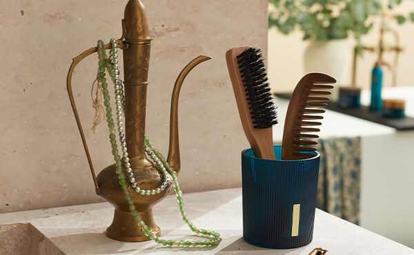 A blue ribbed candle jar upcycled to store a wooden comb and brush, styled beside a brass jug with beads.