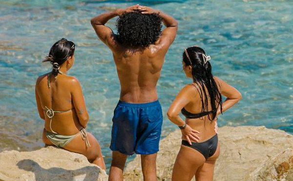 Três amigos em fato de banho a relaxar à beira-mar, desfrutando juntos do sol, da ligação e de momentos de verão despreocupados.