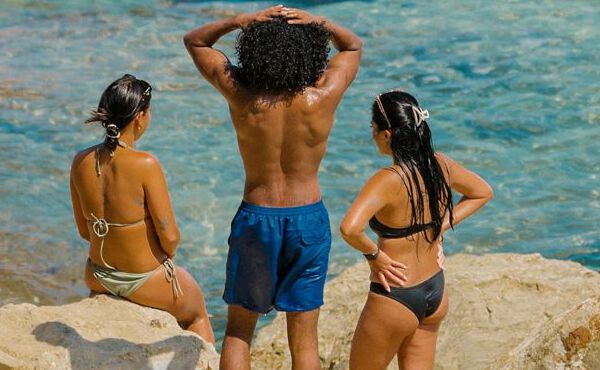 Tres personas se relajan junto al mar vestidas con ropa de baño, disfrutando de la amistad, el sol y la despreocupación del verano.