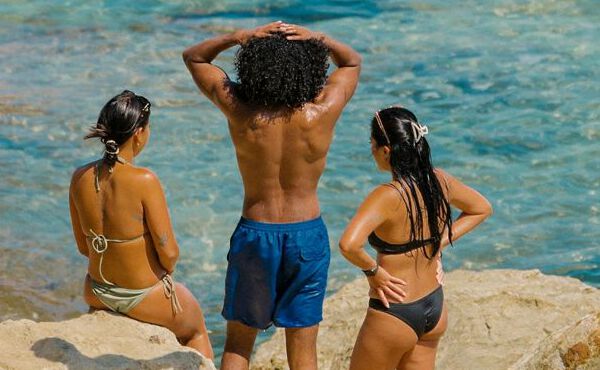 Drei Freunde in Badekleidung, die gemeinsam am Meer entspannen, die Sonne genießen und unbeschwerte Sommermomente erleben.