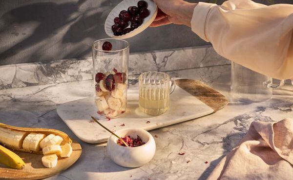 Een persoon die kersen toevoegt aan een glas met bananenschijfjes en citroensap tijdens het bereiden van een smoothie voor een gezonde huid.