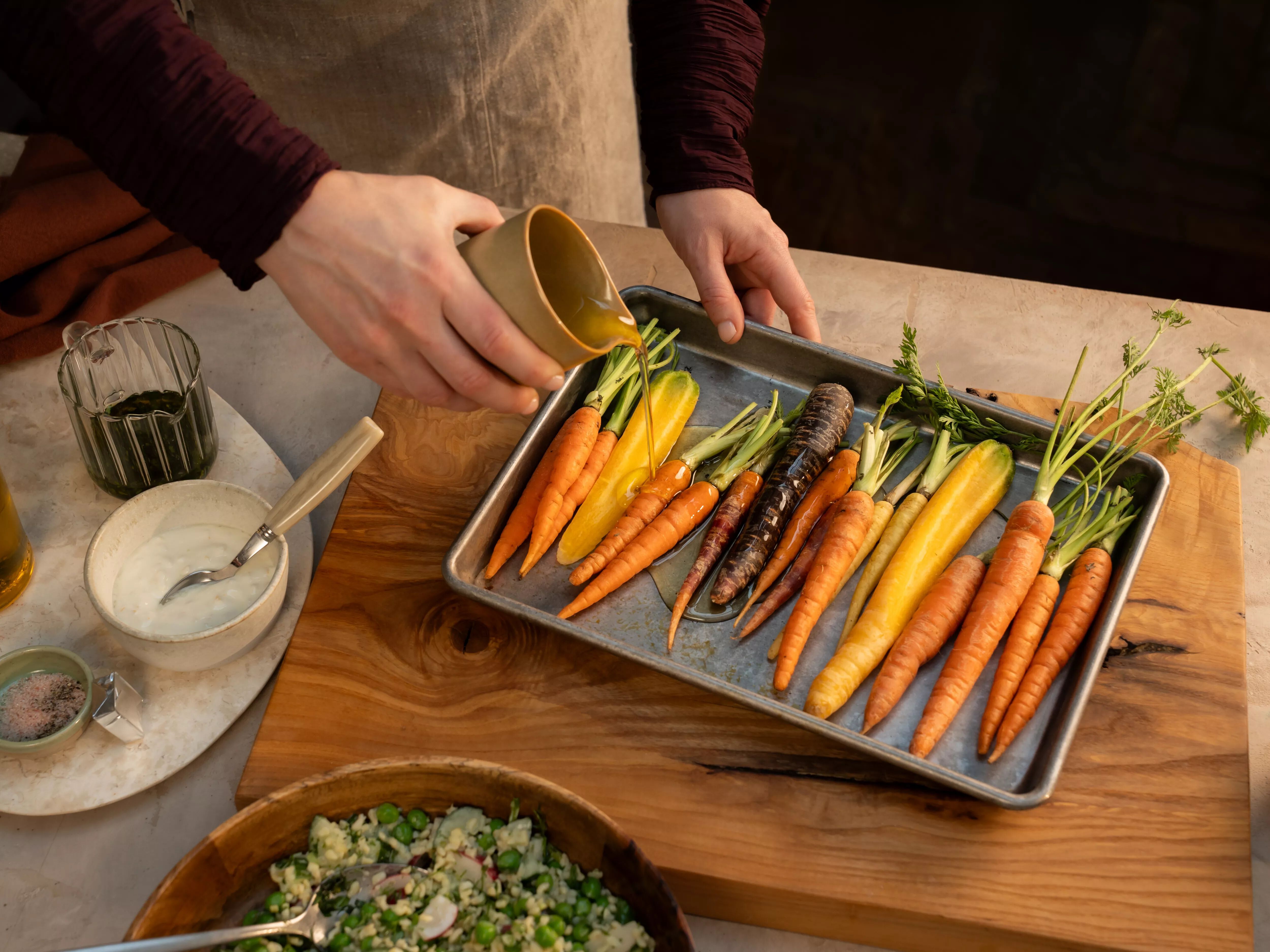 Person drizzling olive oil over a tray of colorful heirloom carrots on a wooden cutting board, surrounded by bowls of dressing, seasonings, and salad. 
