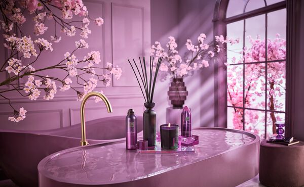 Ein lila Badezimmer mit Kirschbäumen mit rosa Blüten. Es gibt ein großes lila Waschbecken mit goldenem Wasserhahn. Am Waschbecken stehen Produkte aus The Ritual of Yozakura: die Duftstäbchen, der Duschschaum, eine Kerze und die Körpermousse.