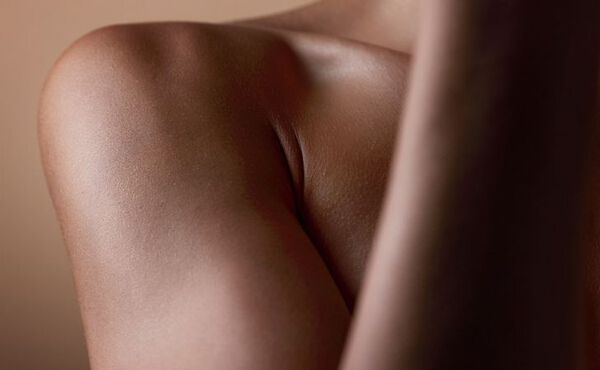 Gros plan sur l’épaule nue et le haut du bras d’une personne avec une peau lisse et bien éclairée, devant un arrière-plan neutre.
