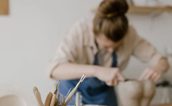 En kvinde former ler, mens der er fokus på nogle keramikværktøjer på et arbejdsbord i forgrunden.