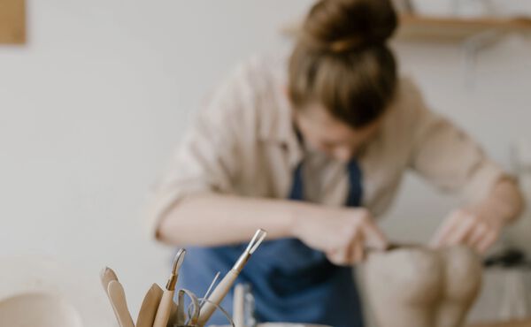 Femme en train de sculpter de l’argile avec des outils de poterie sur une table de travail dans le fond.