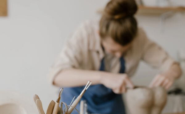 Een vrouw die kleivormen maakt, terwijl op de voorgrond het pottenbakkersgereedschap dat op een werktafel ligt scherp in beeld is.